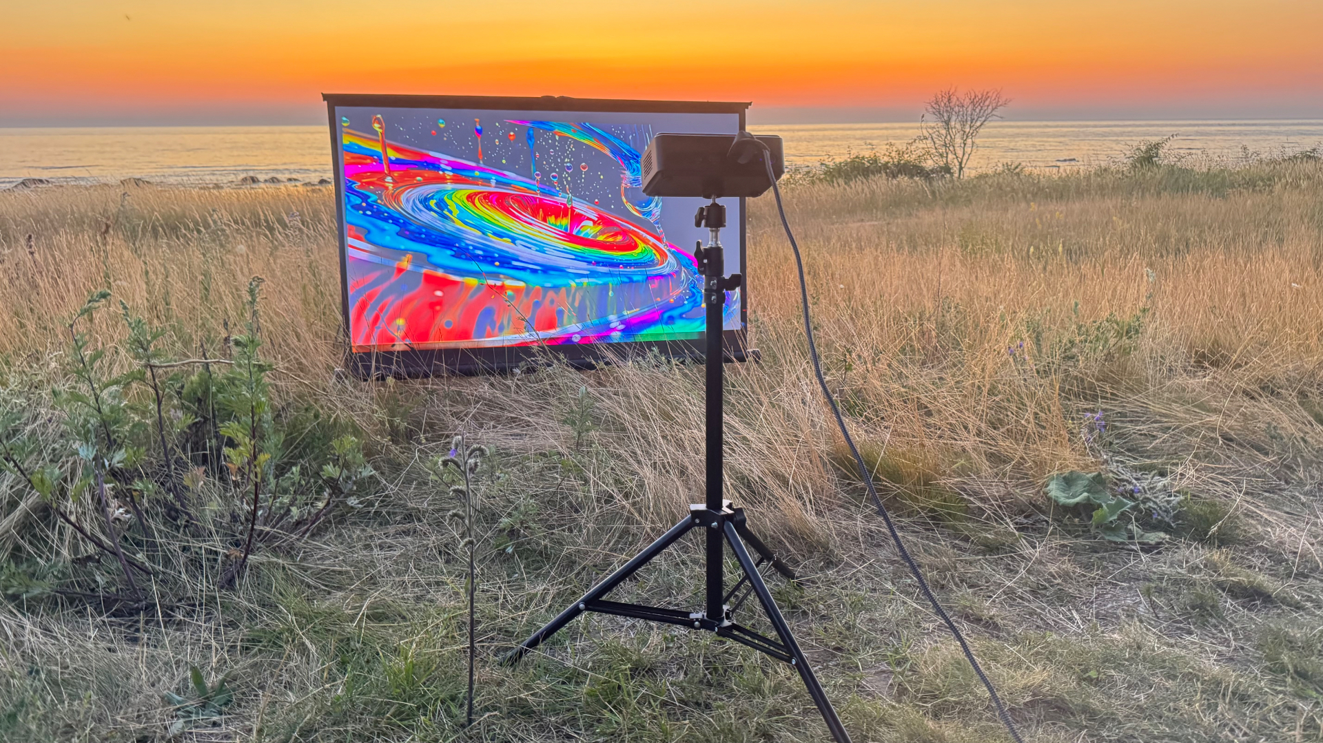 PICZO Nova Ultra S på stativ projicerar färgstark ljuskonst på duk vid havet i solnedgång